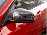 A car with a carbon fiber side mirror is parked outdoors, reflecting nearby buildings in its glossy surface. The car is red and positioned on a street.