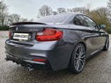 A dark gray BMW M235i is parked on a wet, textured ground with barren trees in the background. The license plate reads 