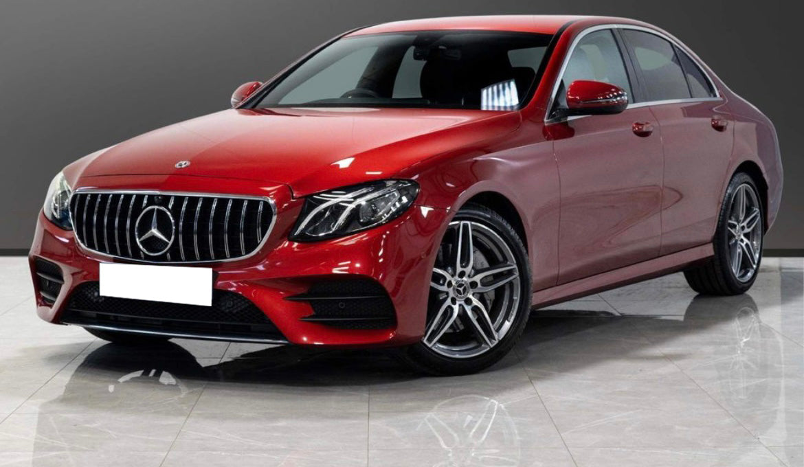 A red Mercedes-Benz sedan is parked on a reflective tiled floor, with a gray gradient background. The car's design features prominent silver rims and a front grille with the iconic emblem.