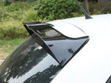A black car spoiler is attached to the roof of a white vehicle, parked outdoors near green bushes and trees, with the word 