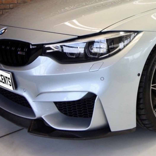 A silver car front, with sleek headlights and black grille, displays a "CARBON ACCENTS" plate. It's positioned indoors against a brick wall backdrop.