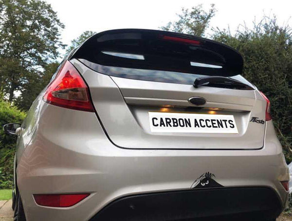 A silver car with a rear spoiler and "CARBON ACCENTS" on the rear plate is parked in a driveway surrounded by trees and bushes.