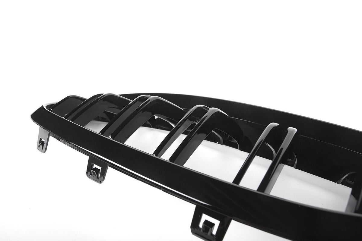 A glossy black car grille with vertical slats rests against a bright white background, featuring attachment clips at the bottom edge.