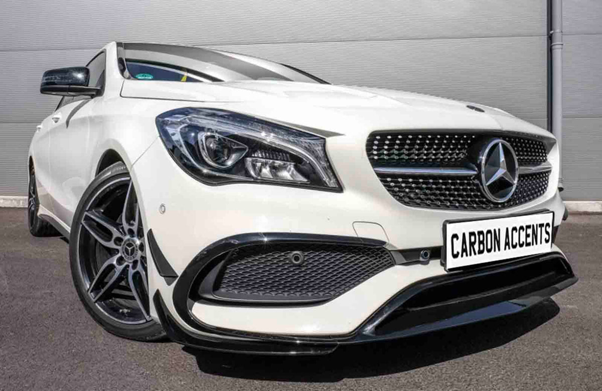 A white car with a Mercedes emblem and sporty design stands stationary on pavement, near a corrugated metal wall. License plate reads "CARBON ACCENTS."