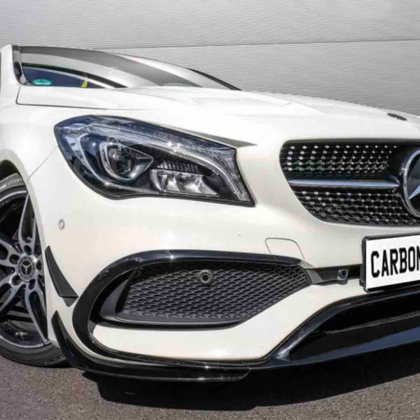 A white car with a Mercedes emblem and sporty design stands stationary on pavement, near a corrugated metal wall. License plate reads "CARBON ACCENTS."