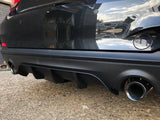 A black car's rear bumper has a visible exhaust pipe and diffuser, reflecting a brick wall and blue sky, parked on a gravel surface.