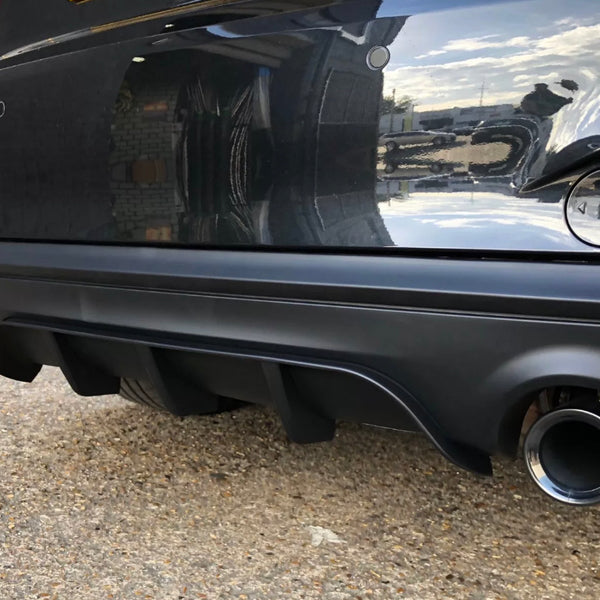 A black car's rear bumper has a visible exhaust pipe and diffuser, reflecting a brick wall and blue sky, parked on a gravel surface.