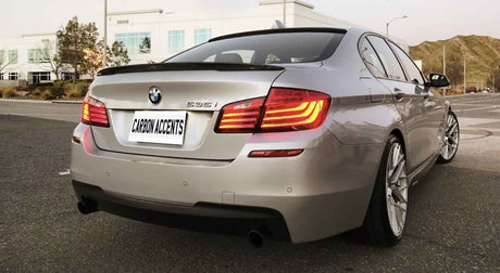 A silver BMW 535i with illuminated brake lights is parked on a road. Surrounding buildings and hills are in the background. The license plate reads "CARBON ACCENTS."