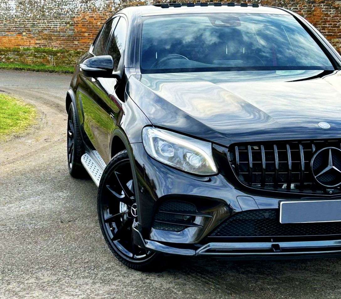 A sleek black SUV, parked on a paved road, features a prominent grille and shiny alloy wheels. It's set against a backdrop of a brick wall and green grass.