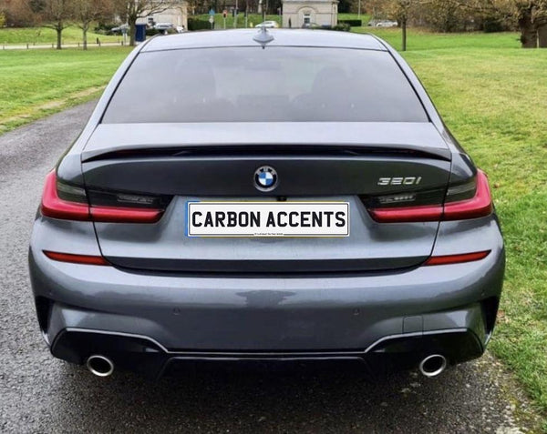 A grey BMW 3 Series car is parked on a paved road, with green grass on either side. The license plate reads "CARBON ACCENTS."