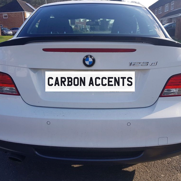 A white BMW 123d is parked in a residential neighborhood. The rear license plate reads "CARBON ACCENTS," with neighboring houses visible in the background.