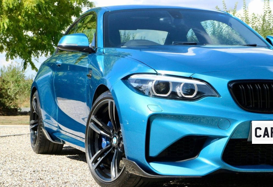 A blue sports car stands on a gravel path, with sleek rims and distinct headlights. Trees and cloudy sky form the background. License plate reads “CAR.”