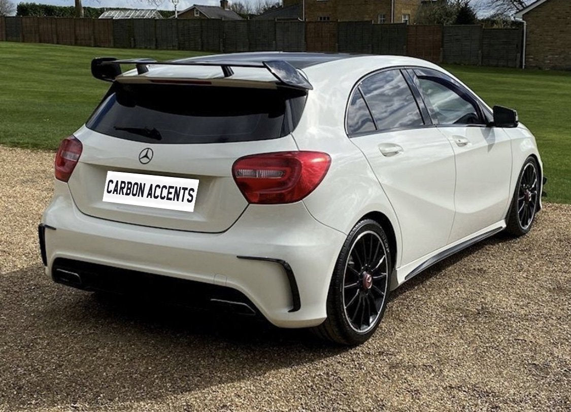 White hatchback car parked on gravel, with black rims and spoiler, viewed from the rear in a grassy area. License plate reads "CARBON ACCENTS."