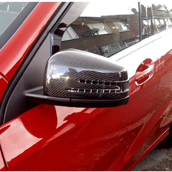 A carbon fiber car's side mirror on a red vehicle reflects a residential building. Nearby windows and building details are visible, indicating an urban street environment.
