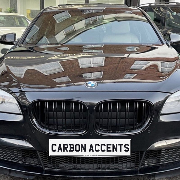 A black BMW is parked, reflecting building windows on its hood. The front license plate reads "CARBON ACCENTS." The car is situated in an urban environment.