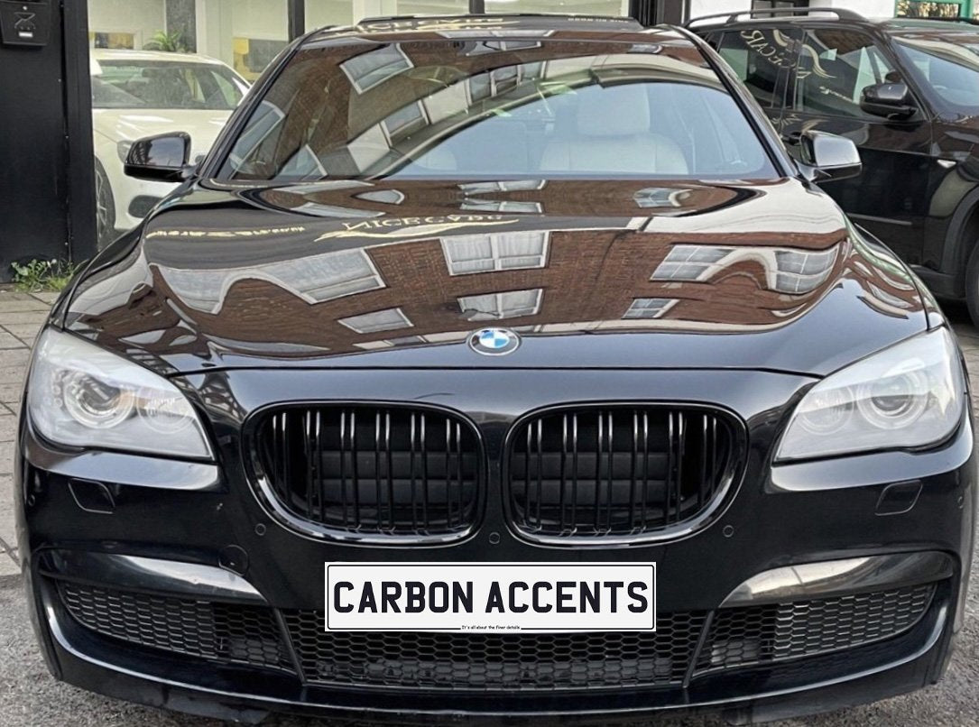 A black BMW is parked, reflecting building windows on its hood. The front license plate reads "CARBON ACCENTS." The car is situated in an urban environment.