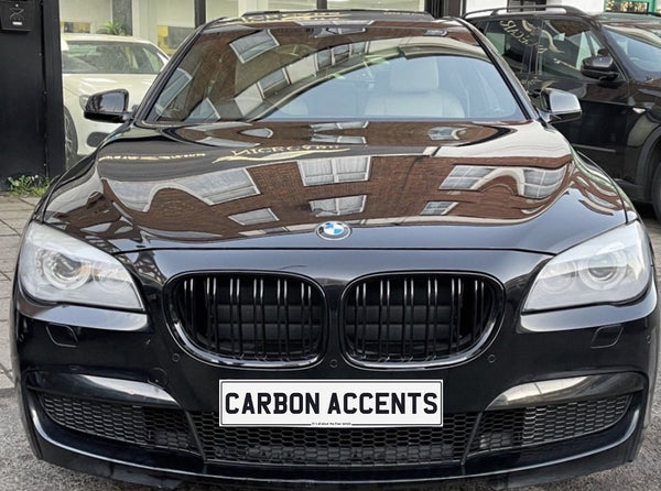 A black BMW is parked, reflecting building windows on its hood. The front license plate reads "CARBON ACCENTS." The car is situated in an urban environment.