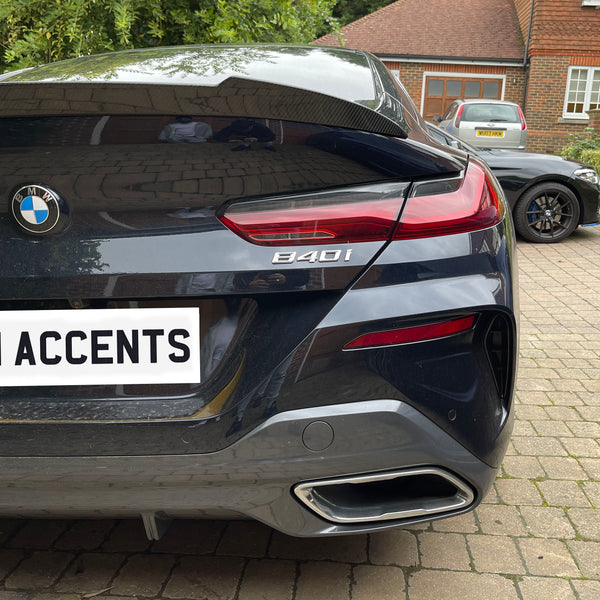 A BMW 840i parked on a brick driveway, displaying its rear badge and tailpipe. Another car is parked nearby. The license plate reads "BN ACCENTS."