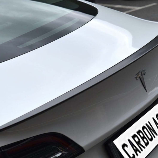 Car trunk with carbon fiber spoiler parked on asphalt, displaying logo and text "CARBON ACCENTS" on license plate. The rear design is sleek and modern.