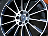 Metallic black and silver car wheel rim with spokes, featuring a three-pointed star logo at the center. The rim rests on a flat, textured surface.