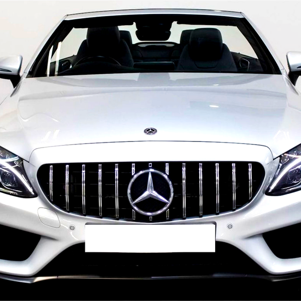 A silver convertible car is stationary in an indoor showroom. It features vertical grille bars, a prominent Mercedes emblem, and sleek, modern headlights. The background is a plain white wall.