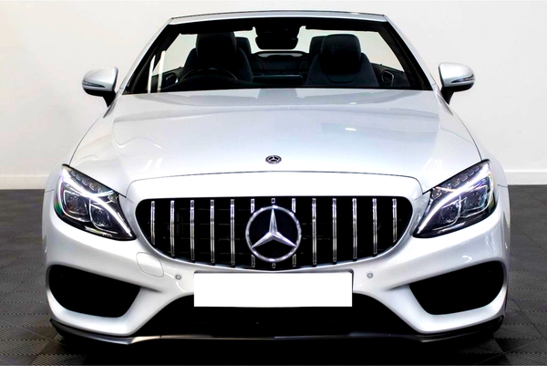 A silver convertible car is stationary in an indoor showroom. It features vertical grille bars, a prominent Mercedes emblem, and sleek, modern headlights. The background is a plain white wall.