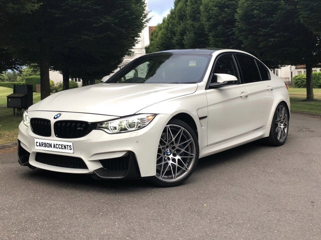 A white BMW car parked on a paved road, featuring silver alloy wheels and "CARBON ACCENTS" on the front plate, surrounded by tall, green trees and grassy areas.