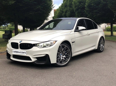 A white BMW car parked on a paved road, featuring silver alloy wheels and "CARBON ACCENTS" on the front plate, surrounded by tall, green trees and grassy areas.