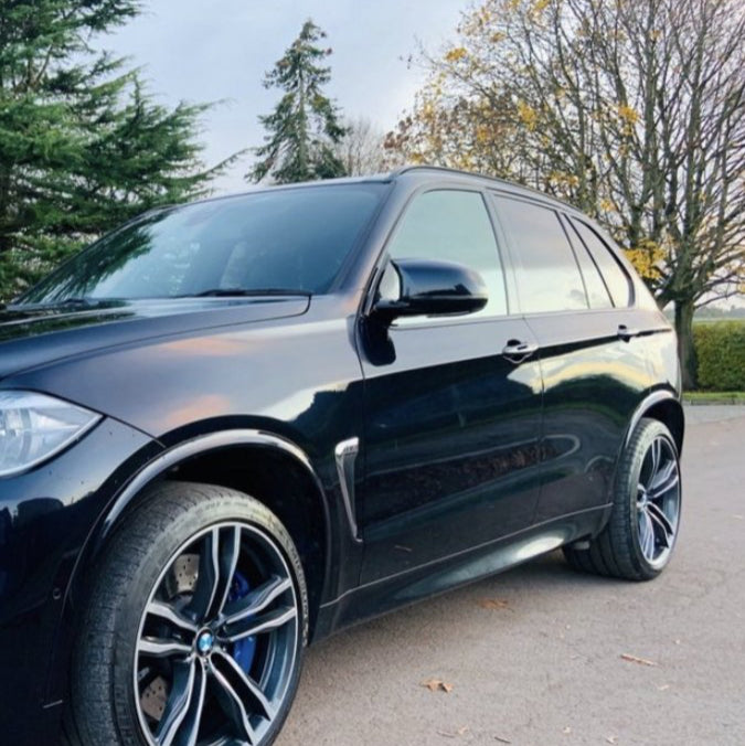 A black SUV is parked on a road, with stylish rims and tinted windows, surrounded by trees with autumn foliage, under a clear sky.