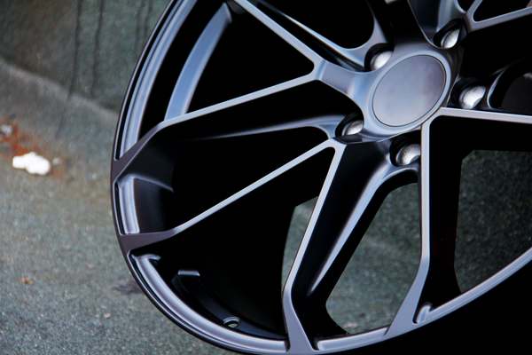 A metallic car wheel rim with a sleek, multi-spoke design rests against a textured asphalt surface, highlighting its dark, polished finish in an urban setting.