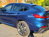 A blue BMW X4 is parked on a residential street, with sunlight highlighting its shiny surface. The license plate reads 