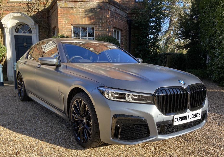 A sleek silver BMW sedan is parked on a gravel driveway in front of a brick house with surrounding greenery. License plate text reads, "CARBON ACCENTS."