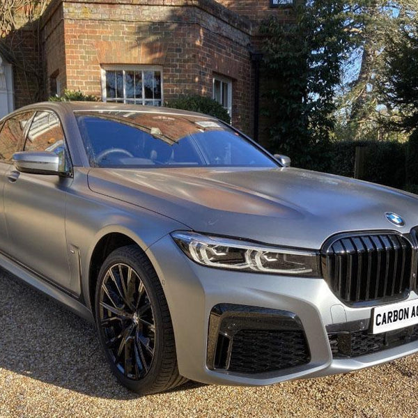 A sleek silver BMW sedan is parked on a gravel driveway in front of a brick house with surrounding greenery. License plate text reads, "CARBON ACCENTS."