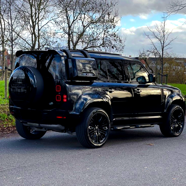 A black SUV is parked on a roadside. It's surrounded by bare trees and green grass, with houses visible in the background under a cloudy sky.