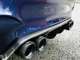Four carbon fiber car exhaust pipes protrude from the rear of a blue vehicle, parked on a textured asphalt surface. A brick wall reflects in the car’s shiny paint.