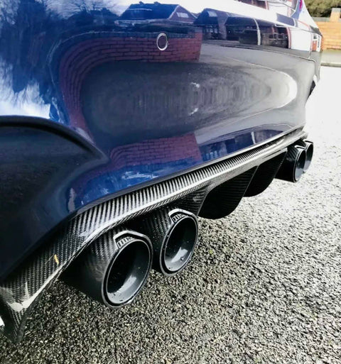 Four carbon fiber car exhaust pipes protrude from the rear of a blue vehicle, parked on a textured asphalt surface. A brick wall reflects in the car’s shiny paint.