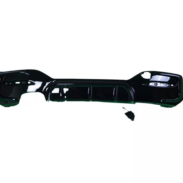 A black car bumper lies flat against a white background. The bumper has several cutouts for attachments, and a small disconnected component rests nearby.