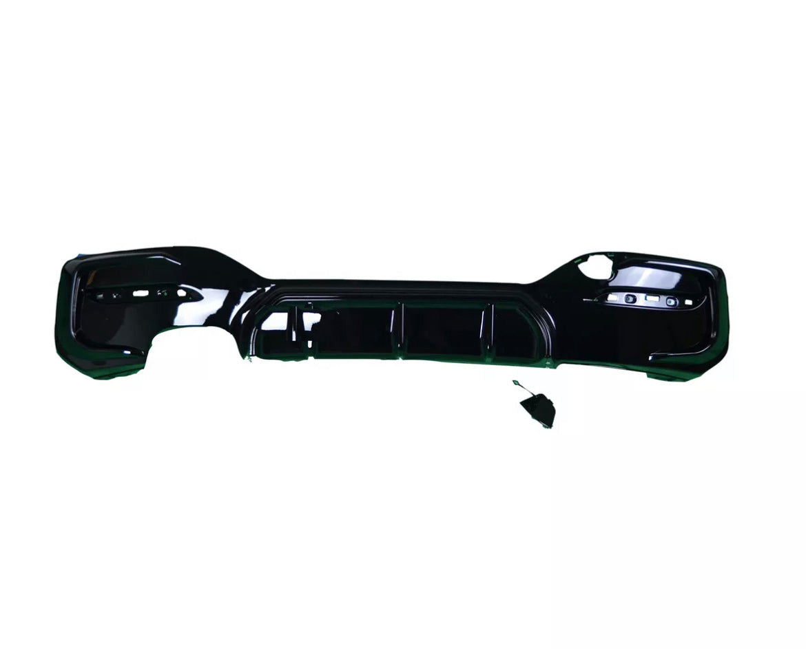 A black car bumper lies flat against a white background. The bumper has several cutouts for attachments, and a small disconnected component rests nearby.