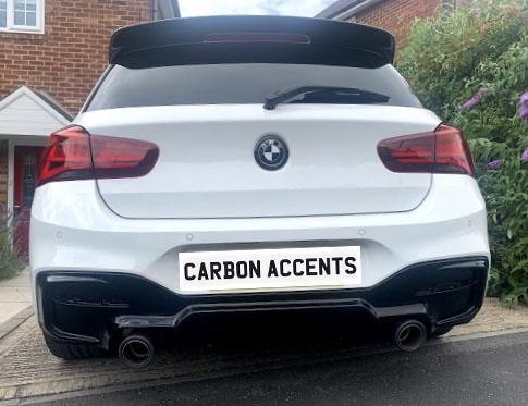 A white car with visible carbon accents is parked on a residential driveway, showing dual exhaust pipes. The license plate reads "CARBON ACCENTS." Brick houses and shrubs are in the background.