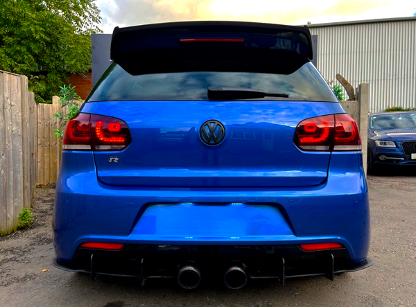 A blue Volkswagen car, parked with visible dual exhausts and a spoiler, is situated between a wooden fence and a metal building, with trees and another car nearby.