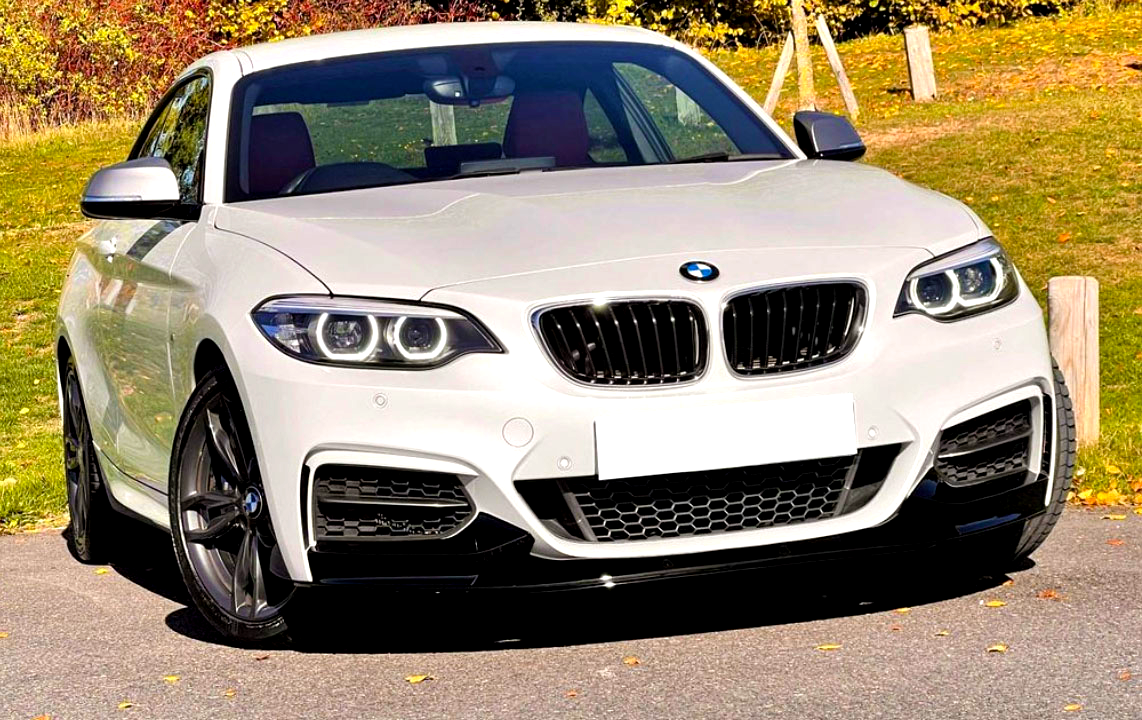 A white BMW car is parked on a paved area, surrounded by green grass and colorful autumn foliage. The front grille and headlights are prominently visible.