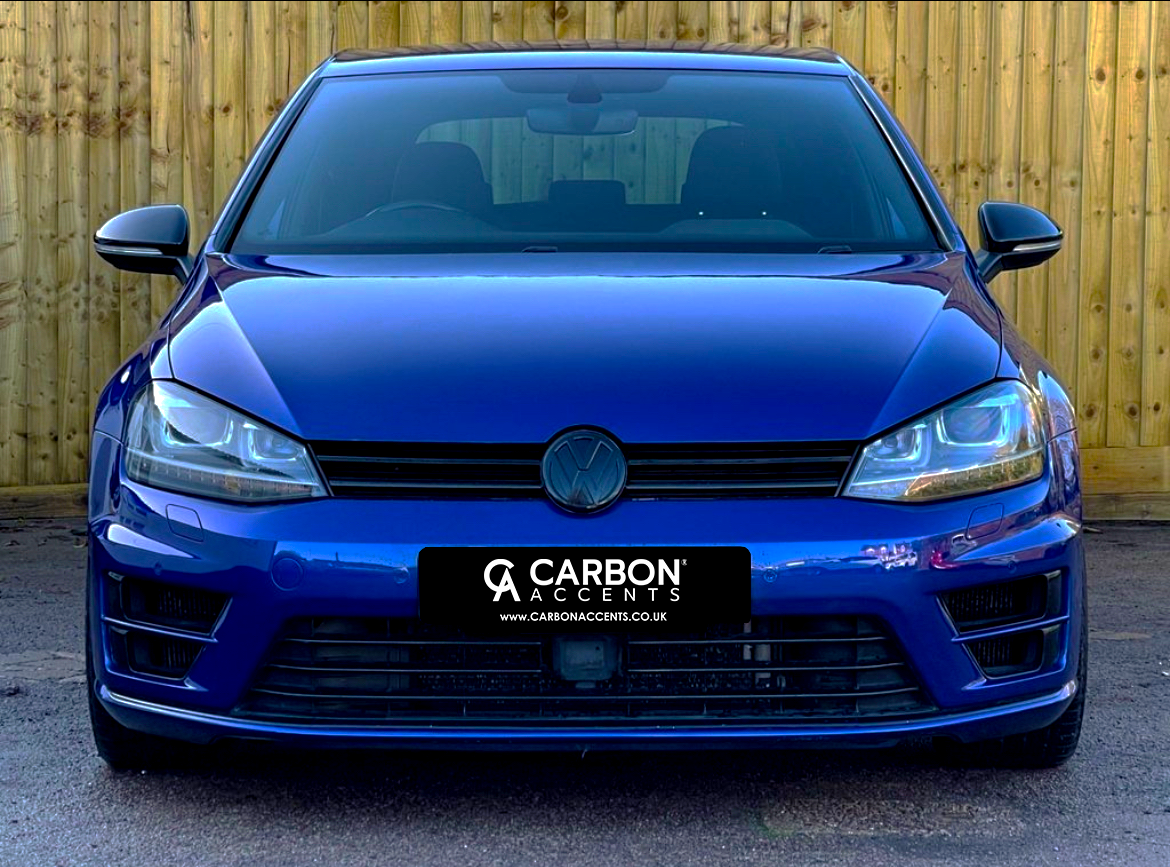 A blue car is parked facing forward, displaying the logo on its grille. "CARBON ACCENTS" is on the license plate. The vehicle is set against a wooden fence background.