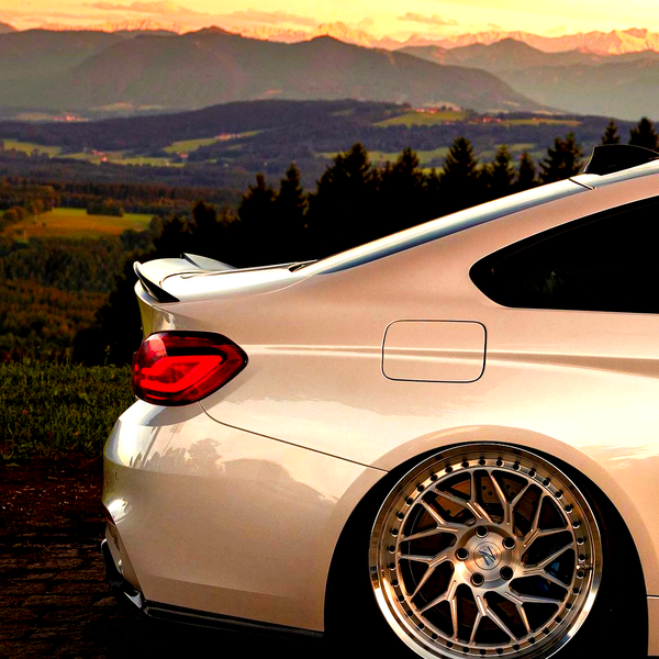 A sleek, silver sports car is parked on cobblestone, showcasing its shiny alloy wheels. It is set against a scenic backdrop of rolling hills and a sunset-lit mountain range.