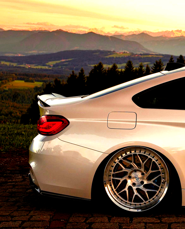 A sleek, silver sports car is parked on cobblestone, showcasing its shiny alloy wheels. It is set against a scenic backdrop of rolling hills and a sunset-lit mountain range.