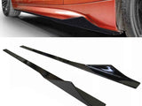 Two glossy black side skirts lie on a white surface, designed for a vehicle shown partially in orange above with one already installed near its tire.