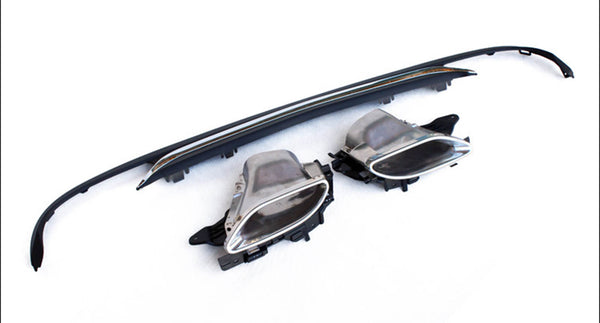 Car exhaust tips and diffuser lie on a white surface, highlighting their metallic and plastic construction. The exhaust tips show a chrome finish, while the diffuser is black and elongated.