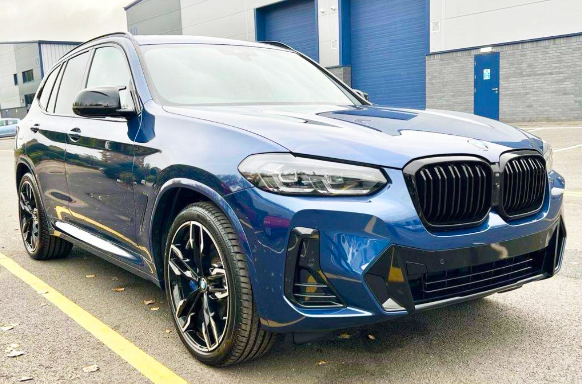 A blue SUV is parked on an asphalt surface with yellow lines, in front of a modern industrial building with large blue doors.