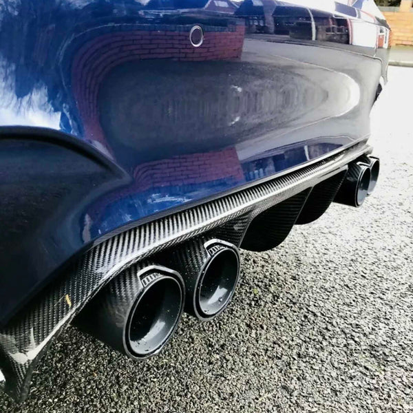 Car exhaust pipes, featuring four round outlets framed by carbon fiber, are situated on a dark car's rear metallic bumper. Parked on a textured asphalt surface, with brick walls reflected.
