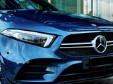A blue Mercedes-Benz car is parked on a cobblestone surface, next to a modern glass building entrance. The focus is on the front grille and headlight details.