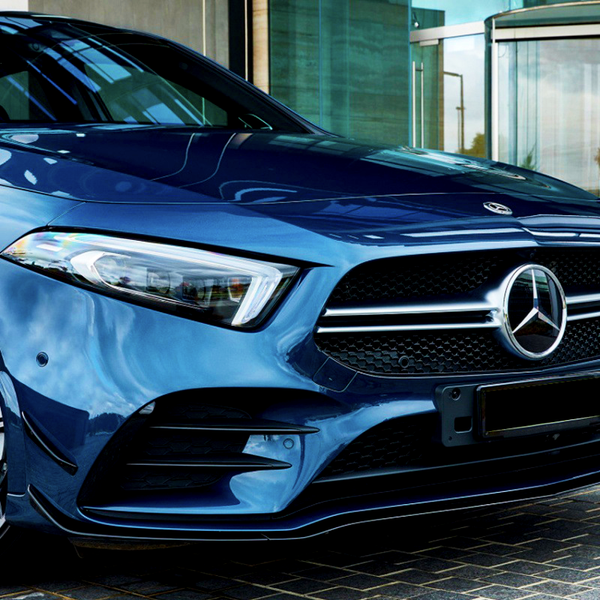A blue Mercedes-Benz car is parked on a cobblestone surface, next to a modern glass building entrance. The focus is on the front grille and headlight details.